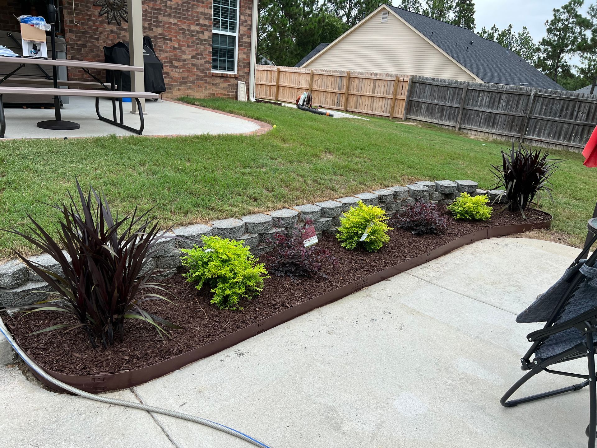 Front yard garden bed with mulch and small shrubs beside a concrete patio and lawn