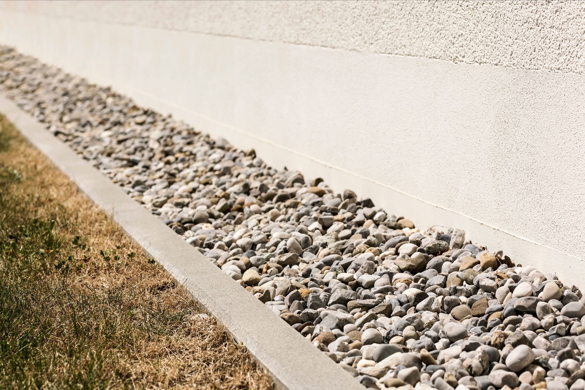 A gravel drainage border sits between a light-colored exterior wall and a patch of dry grass.