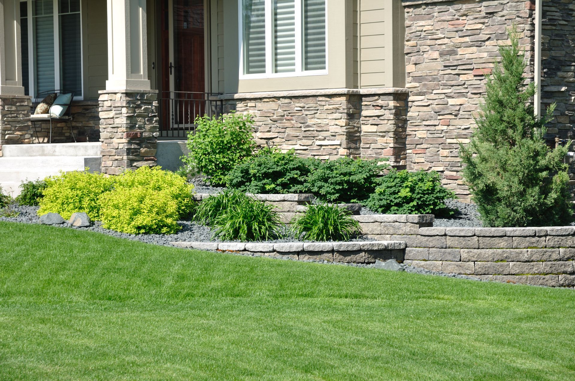 A landscaped front yard features a terraced stone retaining wall, evergreen shrubs, and bright green bushes before a home.