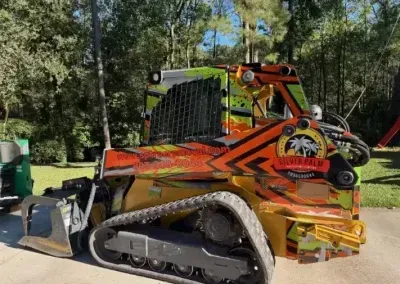 A brightly colored, orange and lime green skid steer loader with a logo on the side, parked on a concrete path outdoors.