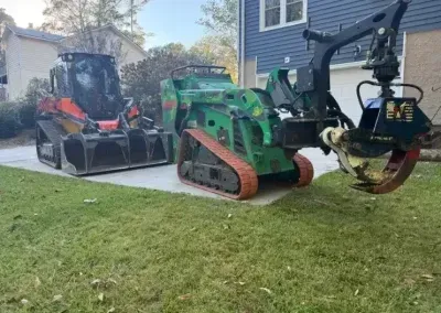 Two tracked skid-steer loaders parked on a residential driveway; one red with a bucket, one green with a tree shear.