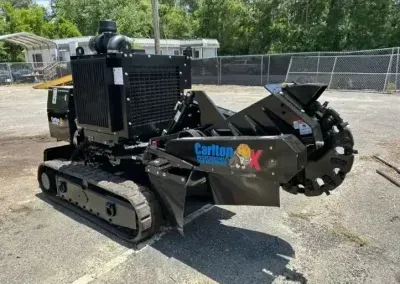 A black Carlton brand tracked trenching machine sits outdoors on a paved lot in front of a chain-link fence.