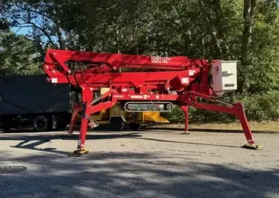 A red track-mounted spider lift stands on pavement with its stabilizers extended in a wooded setting.