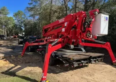 A bright red towable boom lift with extended stabilizing outriggers parked on a dirt path in a wooded area.