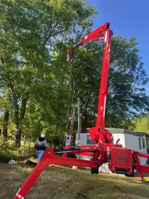 A bright red spider lift crane with an extended boom arm is positioned on a grassy lawn near a house and trees.