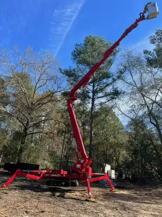 A bright red tracked spider lift extended high against a blue sky, standing on outriggers in a wooded, dirt-covered area.