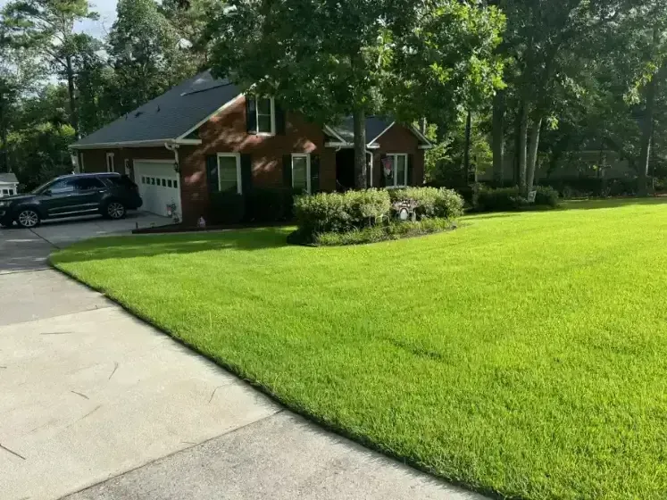 A brick house with a dark car in the driveway and a neatly manicured lawn under large green trees.