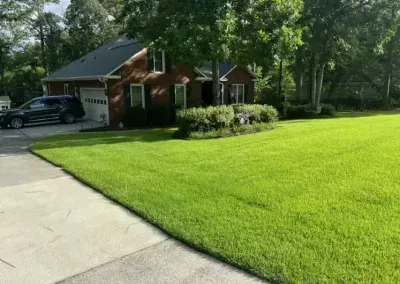 A brick house with a dark car parked on a driveway next to a well-maintained, vibrant green lawn under sunny skies.