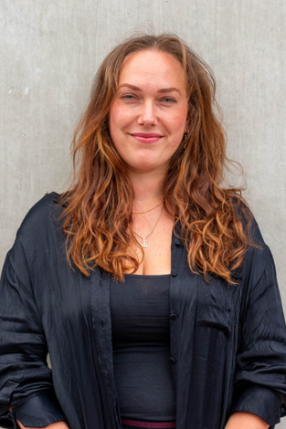 Een vrouw met krullend bruin haar glimlacht voor een grijze muur, gekleed in een zwart shirt en topje.