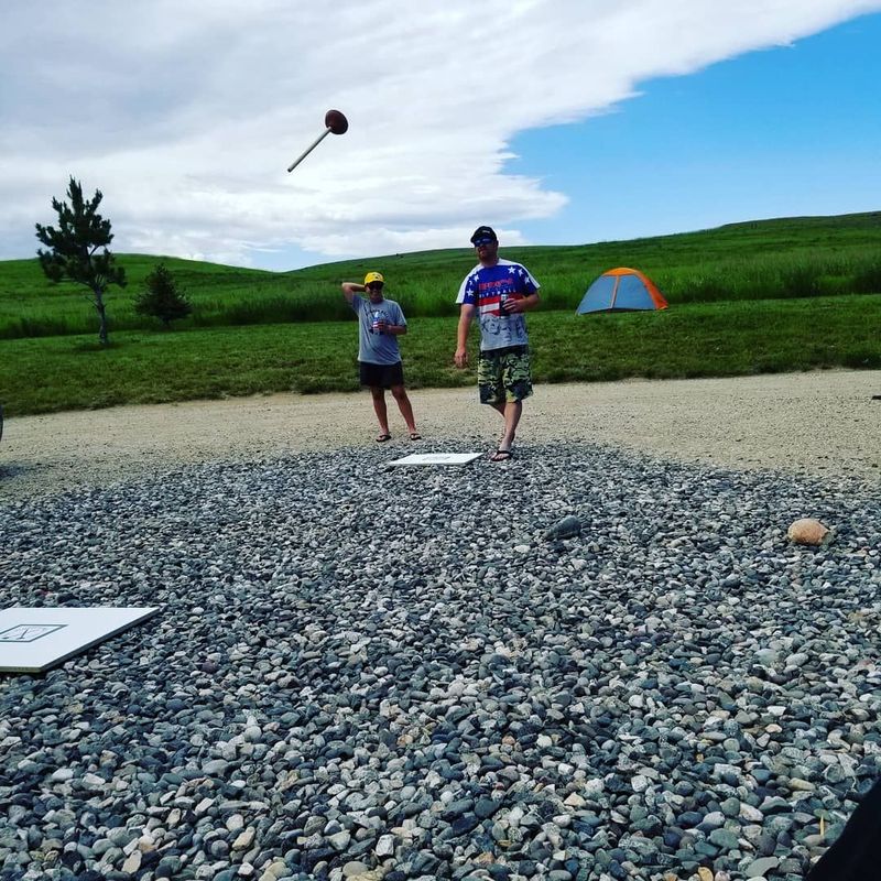 Two people play a ring toss game on a gravel court outdoors with a tent visible in the background.