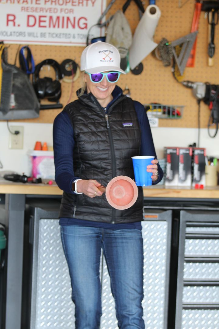 A person in a garage wears a vest, cap, and sunglasses while holding a blue cup and a small bowl.