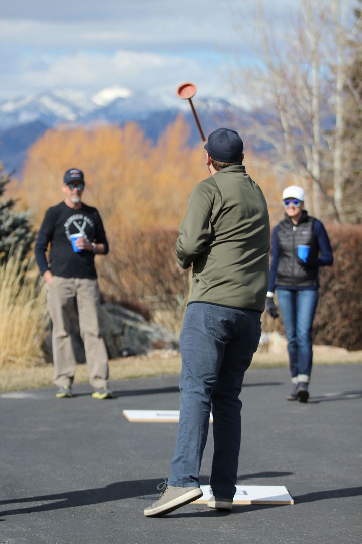 A person throws a plunger toward a target on a paved driveway with two onlookers, mountains, and fall trees in the back.