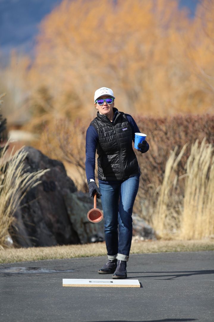 A person in a vest, jeans, cap, and sunglasses stands on a path holding a blue cup and a ball scoop.