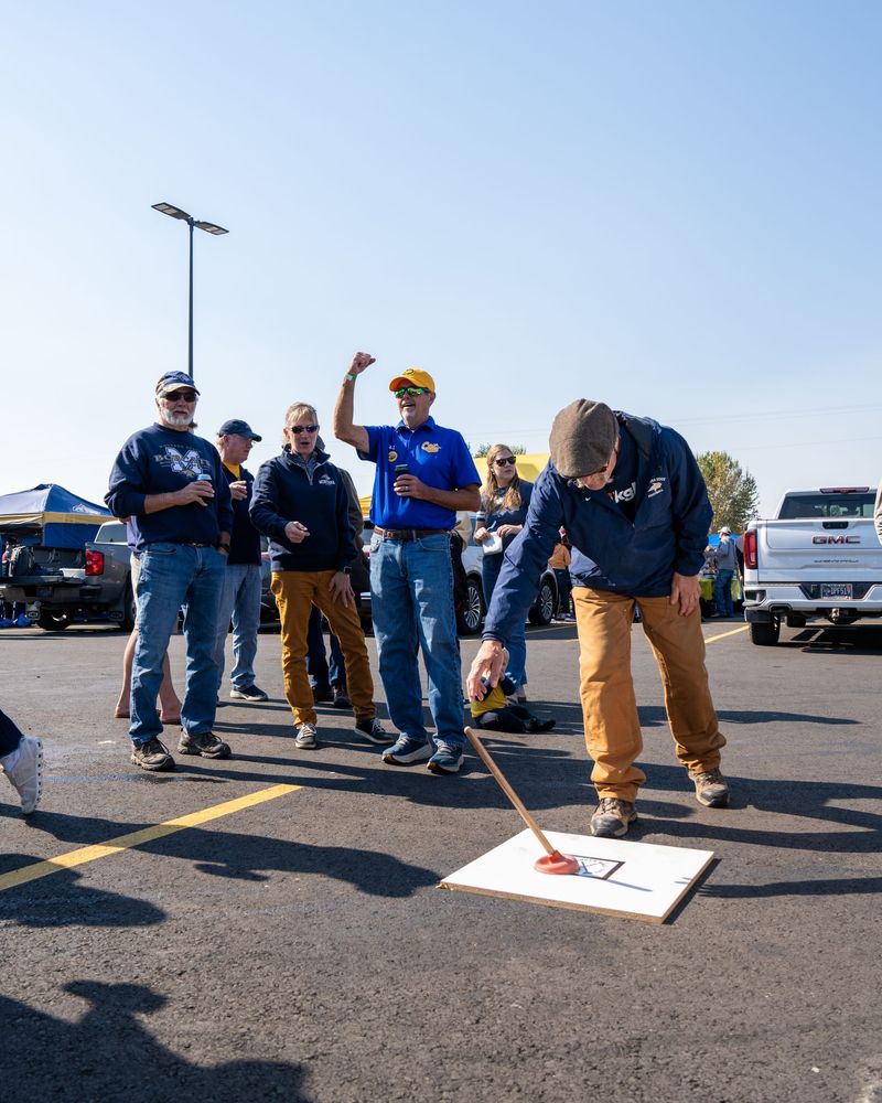 A person uses a plunger to pick up a small item from a board in a parking lot while others watch and cheer.