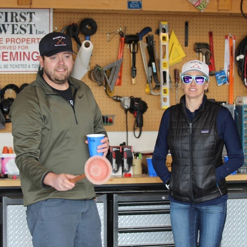 Two people stand in a workshop holding a toilet plunger and a blue cup, with tools mounted on a pegboard background.