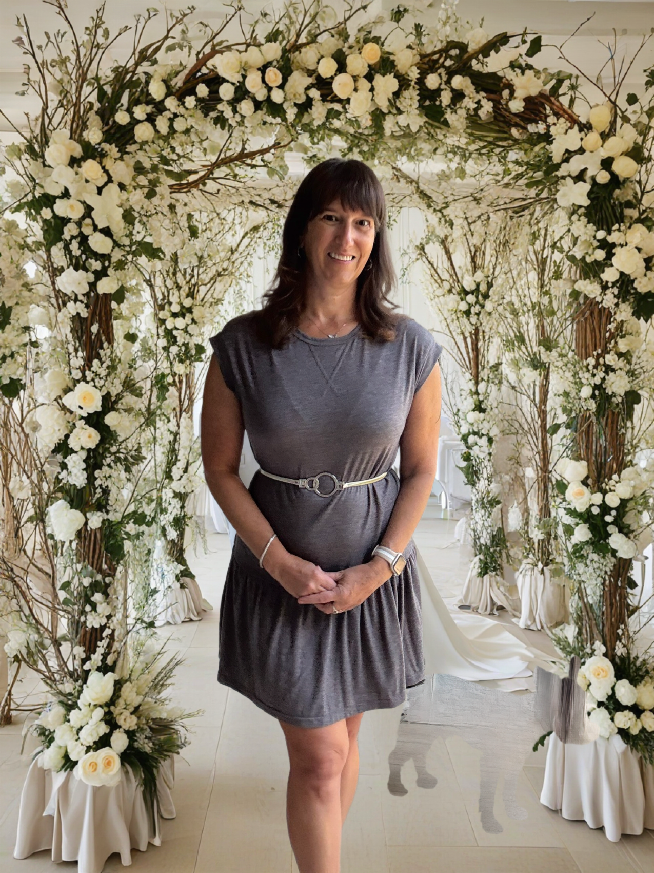 Woman in gray dress stands before floral archway, smiling.