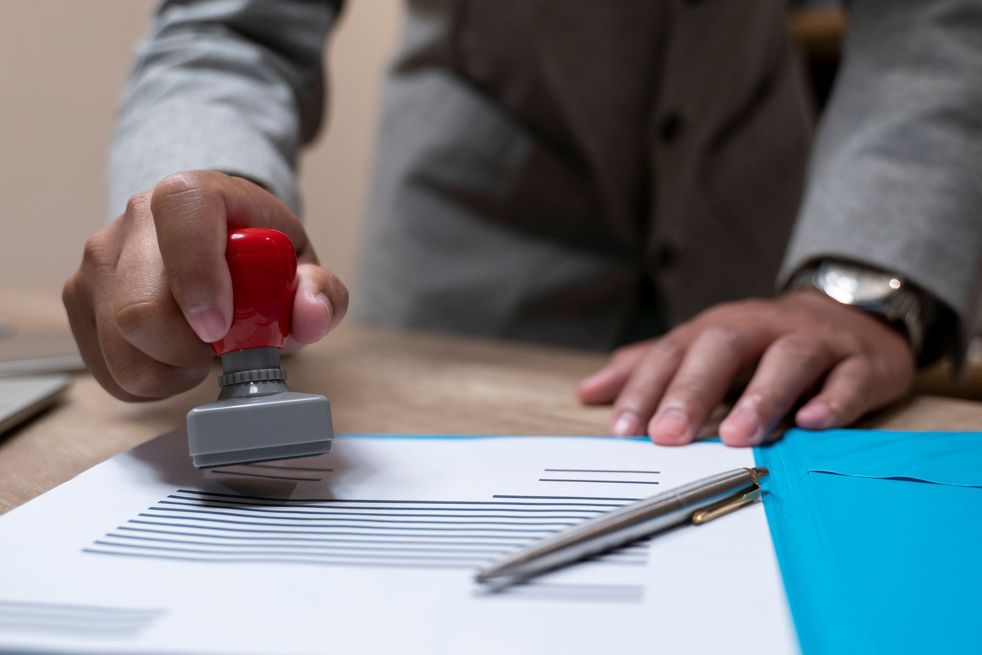 Person in gray suit stamps document with red-handled stamp; pen and blue folder on desk.