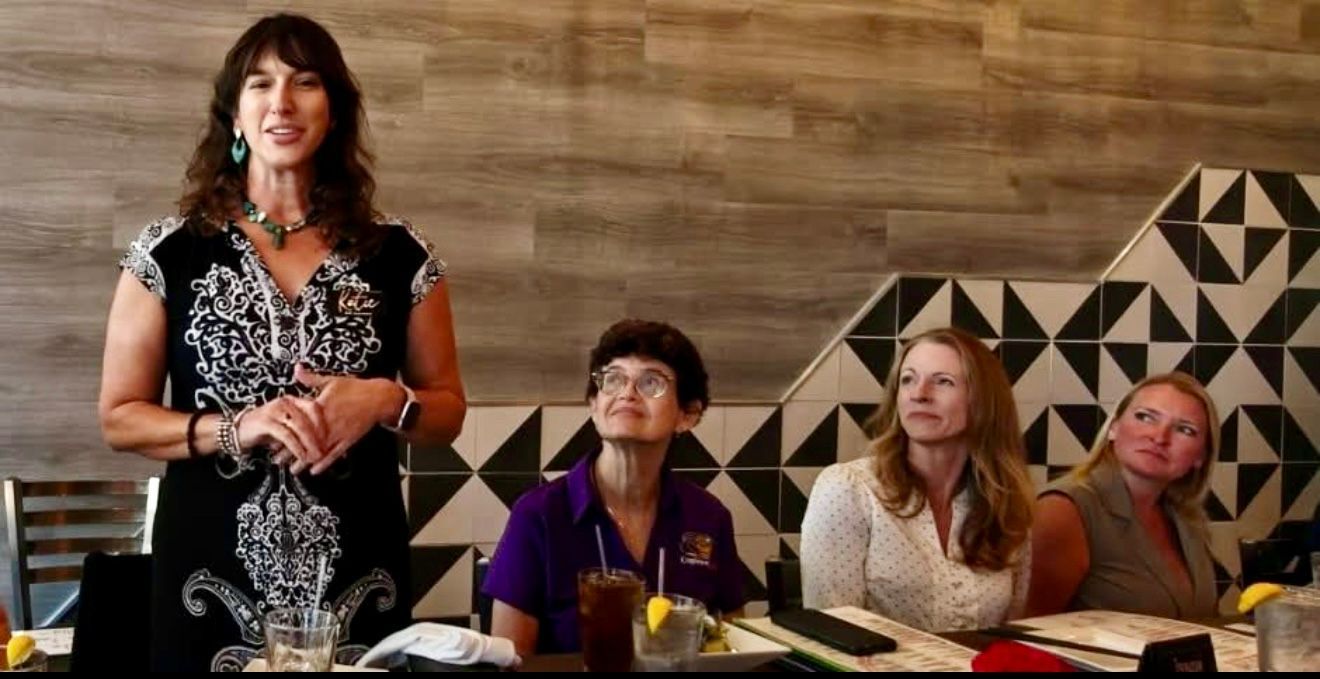 Woman speaking at a table with other women, indoors. Black and white patterned dress. Light brown wood paneling, decorative tile.