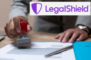 Person in gray suit stamps document with red-handled stamp; pen and blue folder on desk.