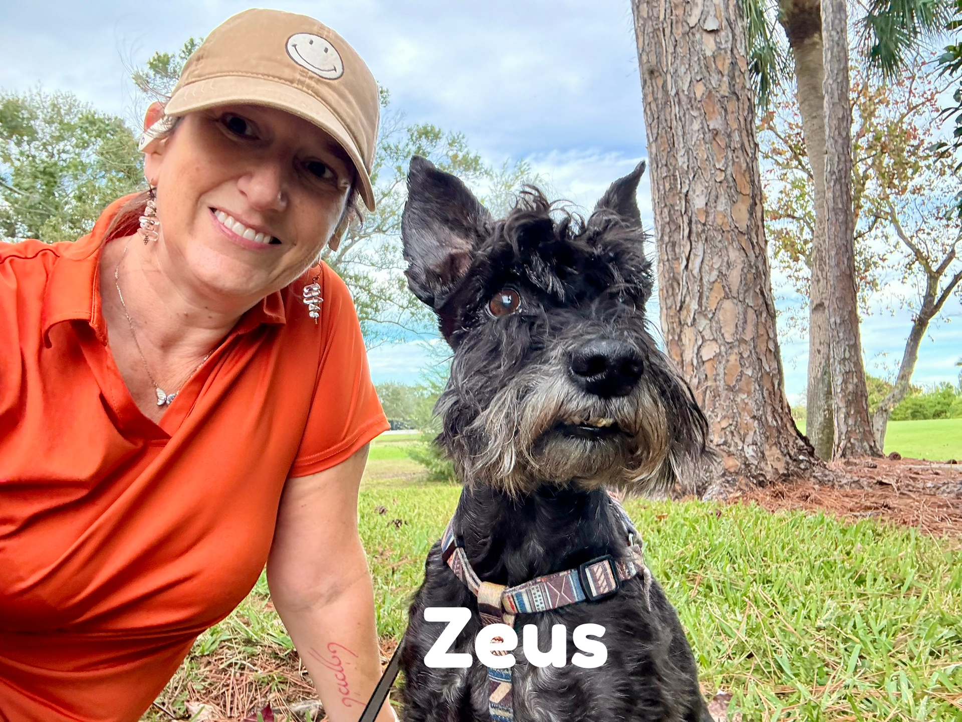 Woman in orange shirt and tan hat smiles next to a black Schnauzer dog named Zeus in an outdoor setting.