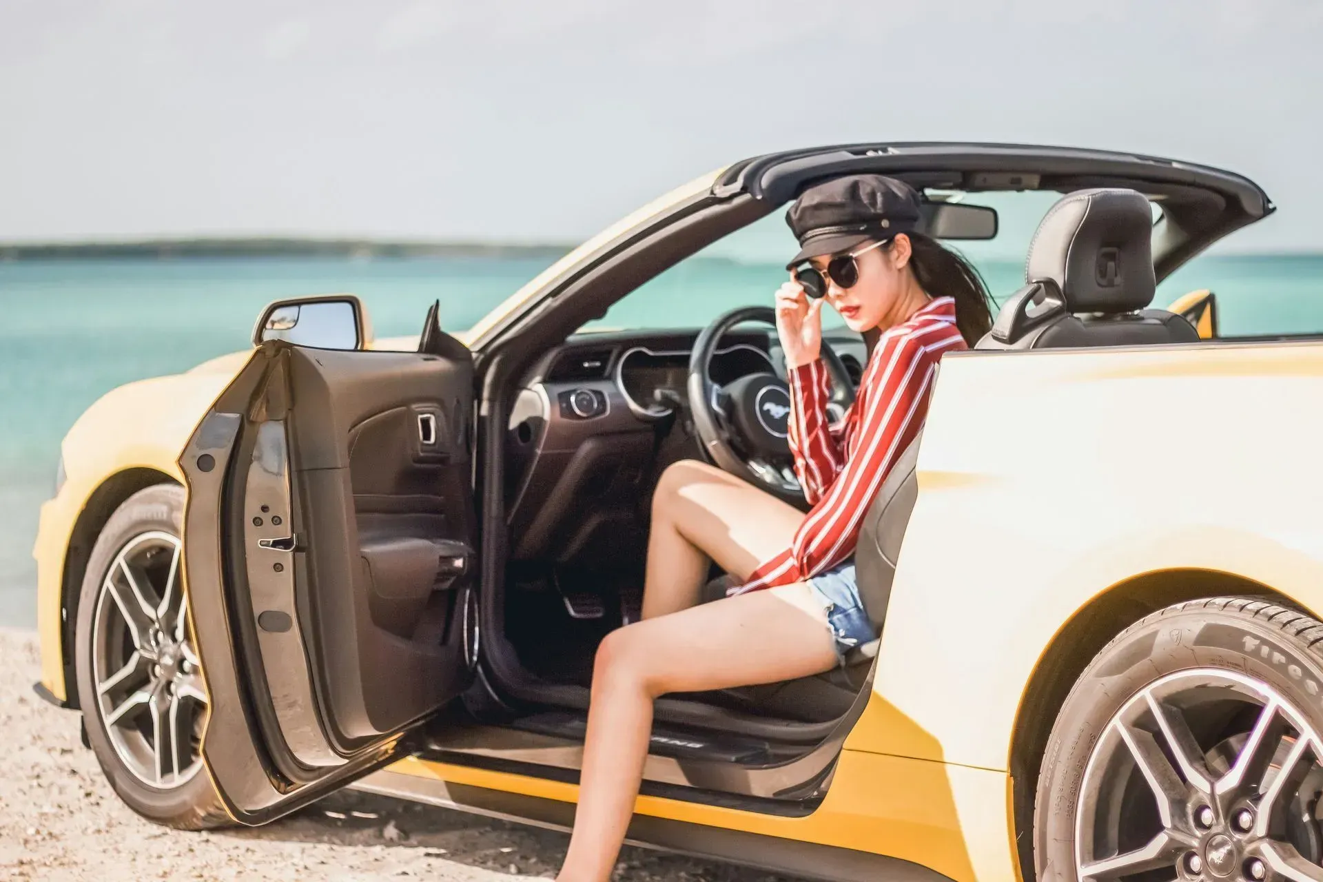 Woman in sunglasses, red striped shirt, and jean shorts sits in a yellow convertible car with the door open; beach in background.