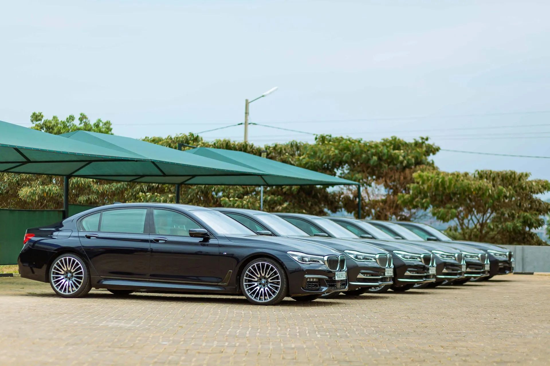 A line of dark luxury sedans parked under a canopy. The setting appears to be outdoors on a sunny day.