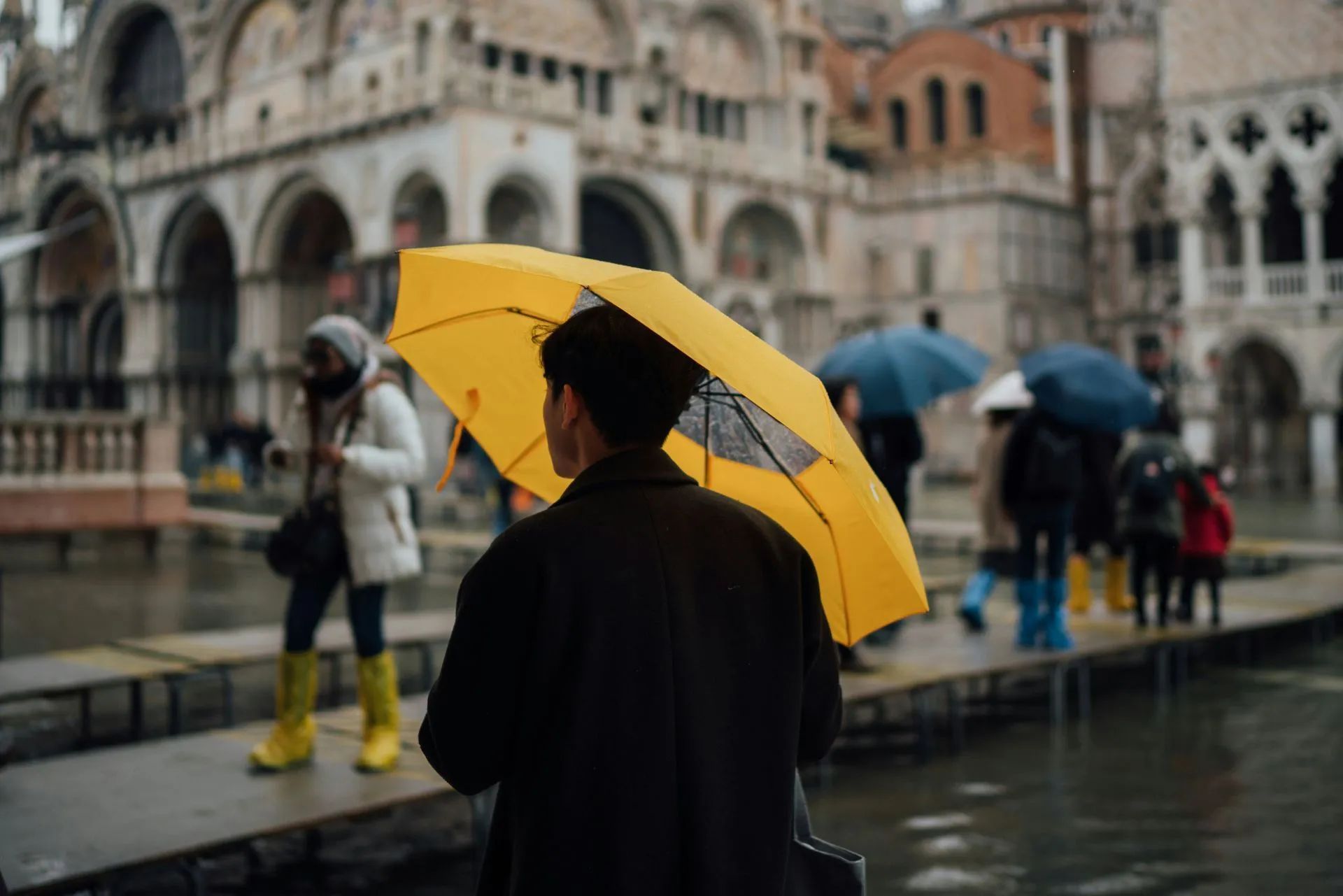Person with yellow umbrella in Venice, Italy, on a rainy day. Other people in rain gear walking near ornate buildings.