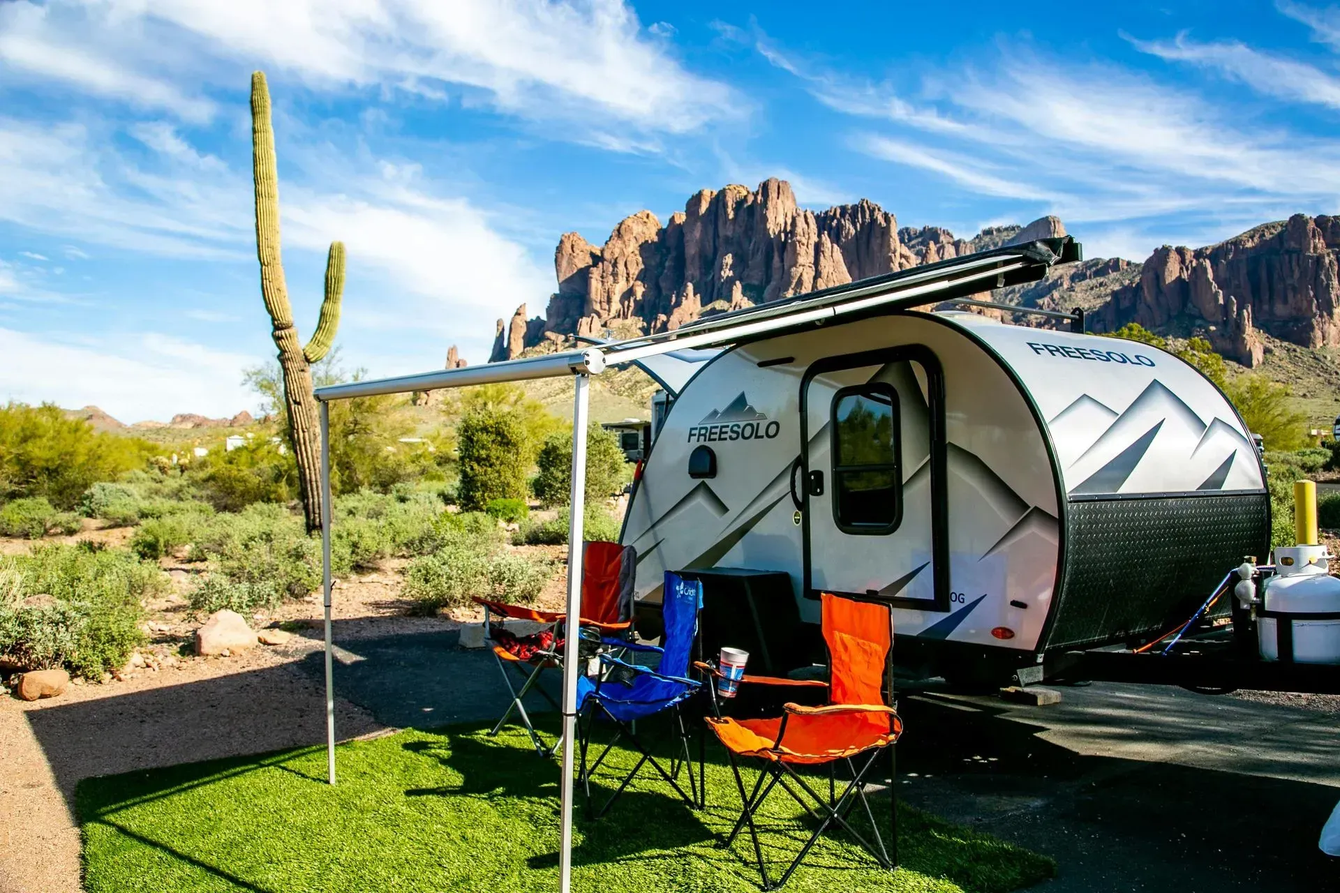 Teardrop camper with awning and folding chairs set up at a campsite, desert landscape in background.