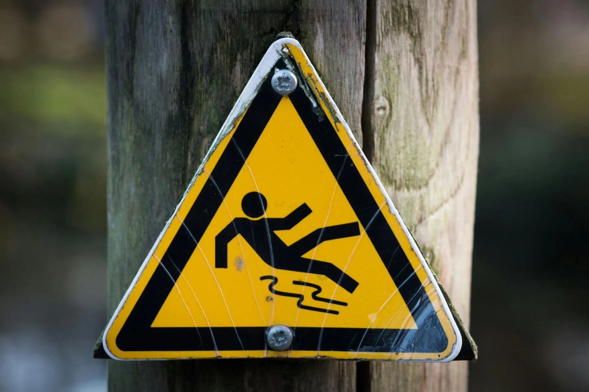 Yellow triangle sign on wooden post warns of a slippery surface with a stick figure slipping.