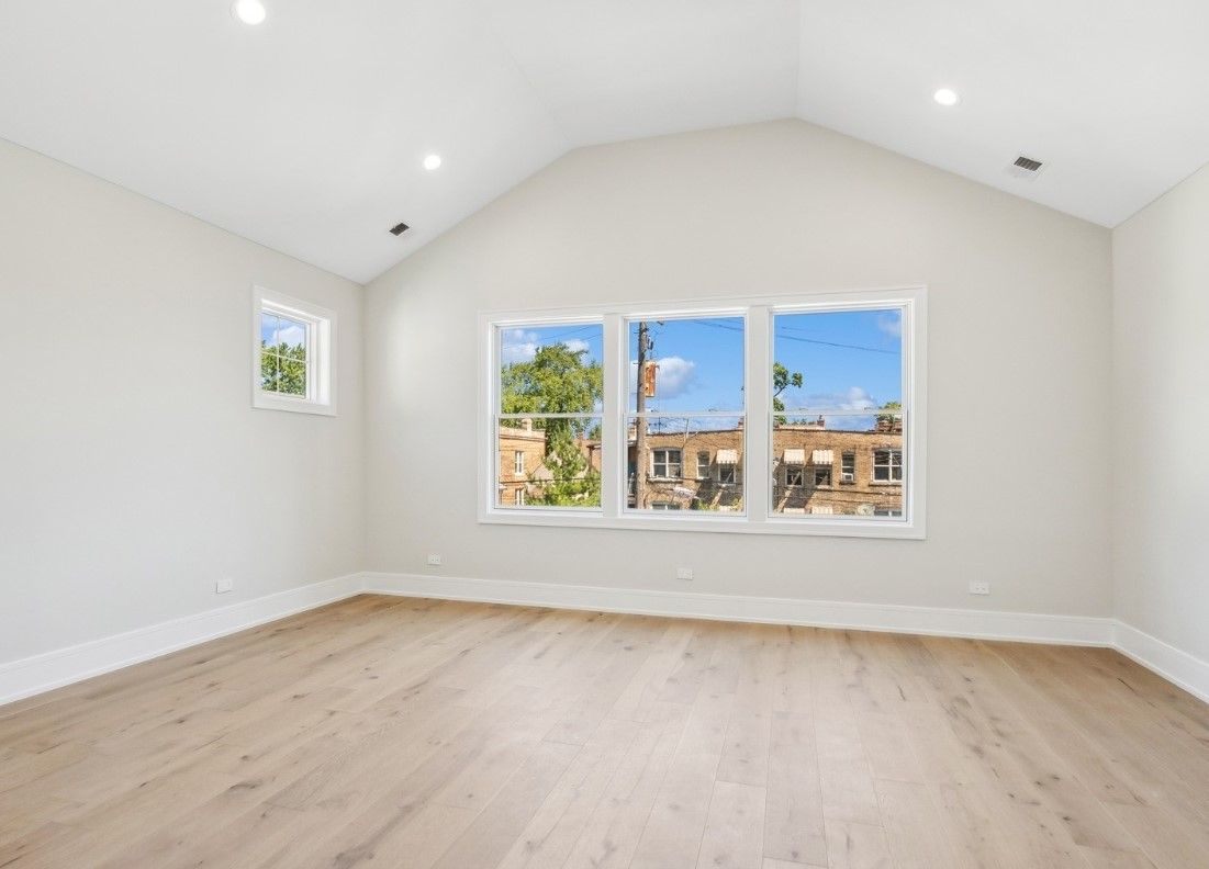 Empty bright room with white walls, vaulted ceiling, light wood floors, and large windows with a city view