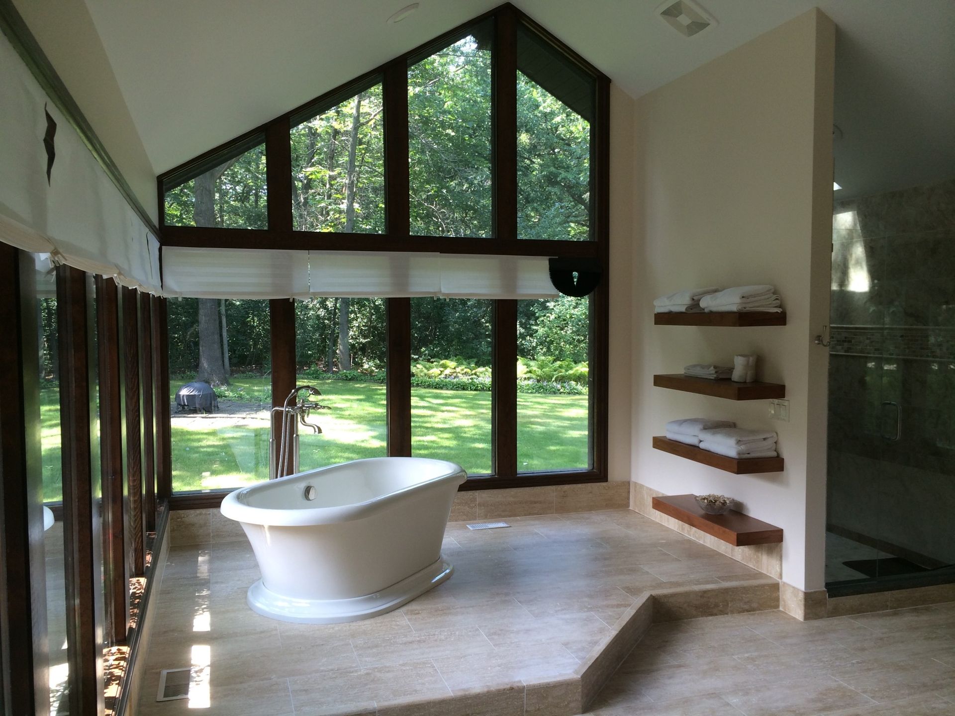 Bathroom with a freestanding bathtub in front of large windows overlooking a green yard, stone floor, and floating shelves.