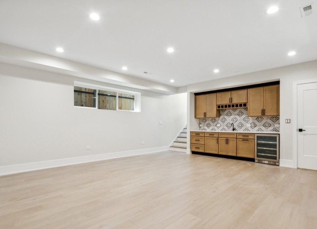 Empty modern basement room with light wood floors, white walls, recessed lights, and a small kitchenette area