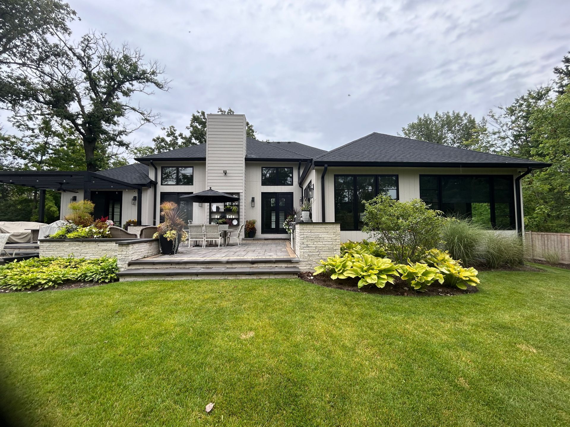 Backyard of a modern house with a patio, lawn, and surrounding greenery under an overcast sky.