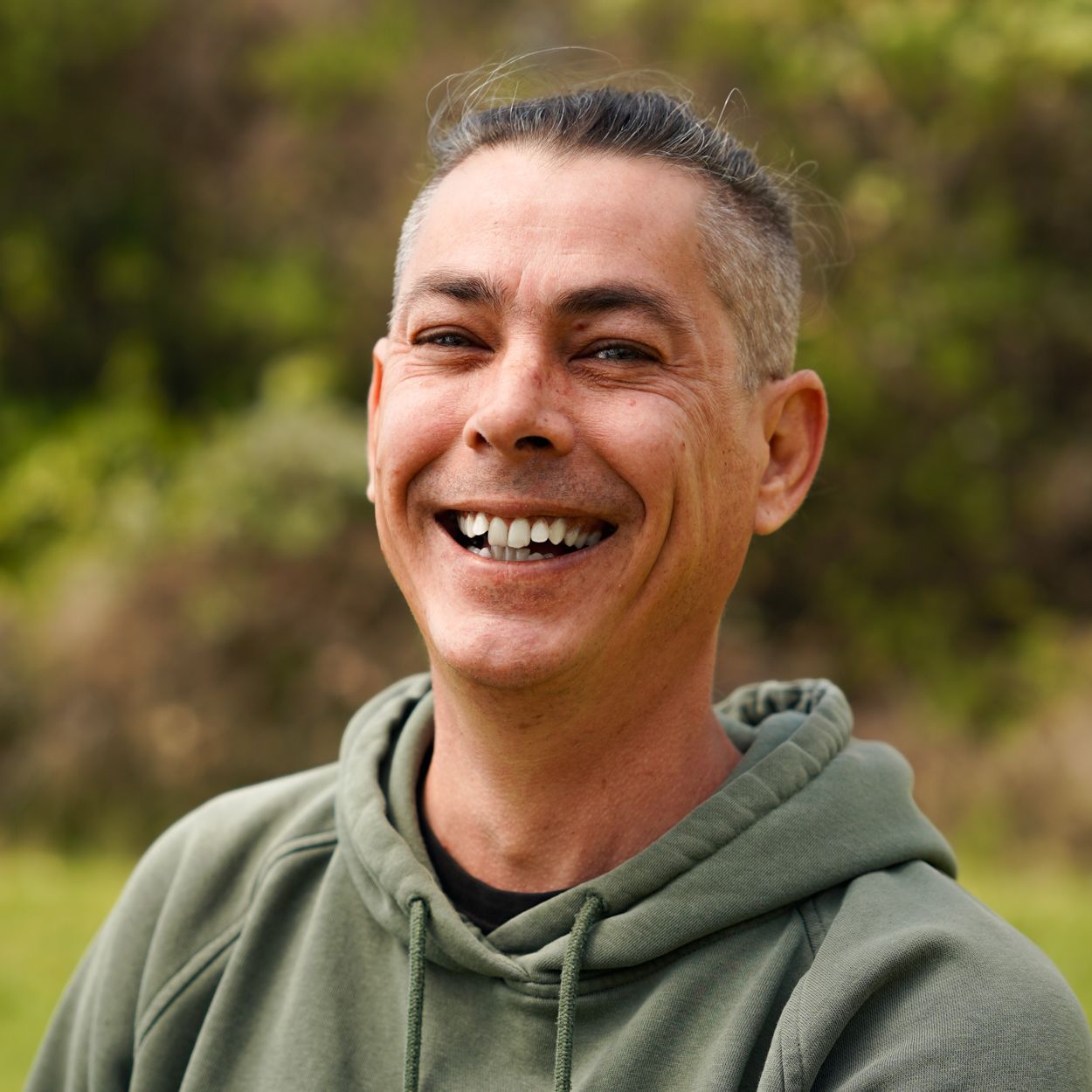 Man with graying hair smiles widely in an olive green hoodie outdoors.