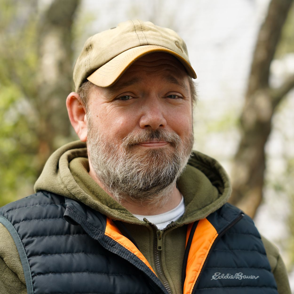 Man with beard, wearing a hat and vest, smiles outdoors.