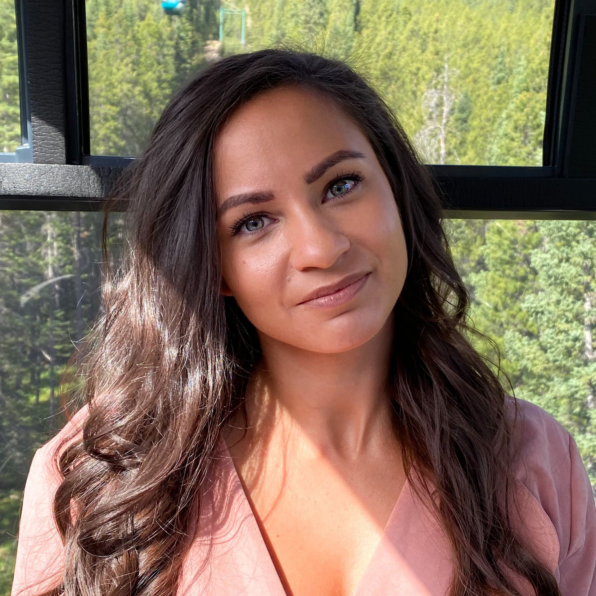 Woman with brown hair and blue eyes, smiling, in a cable car with a mountain view.