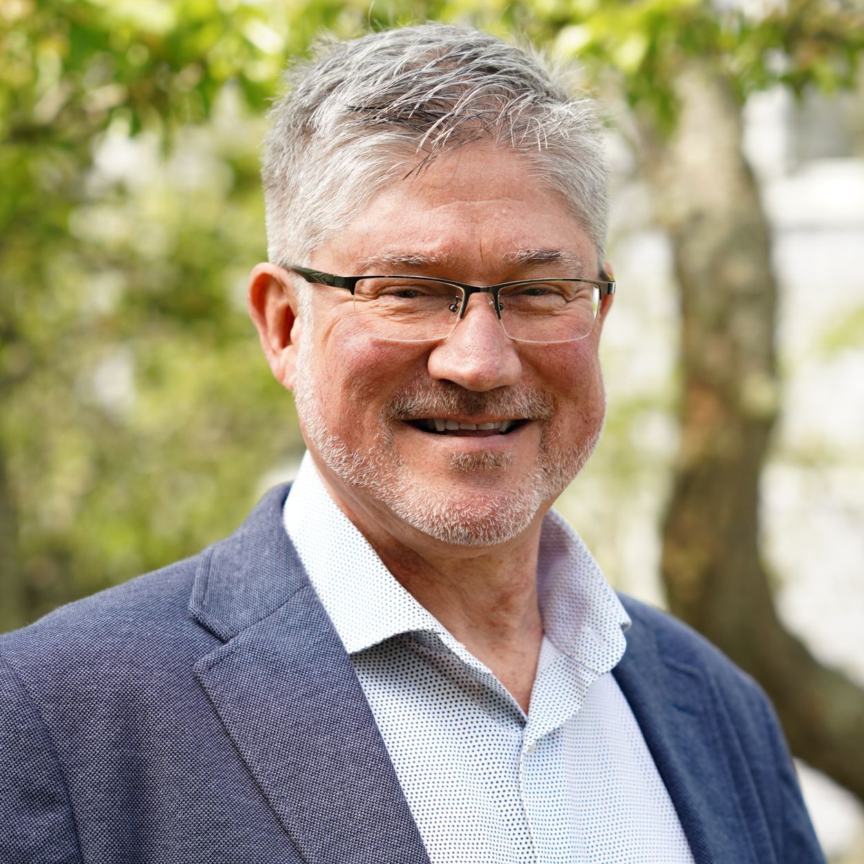 Man with gray hair and glasses smiles, wearing a blue blazer and patterned shirt, outdoors.