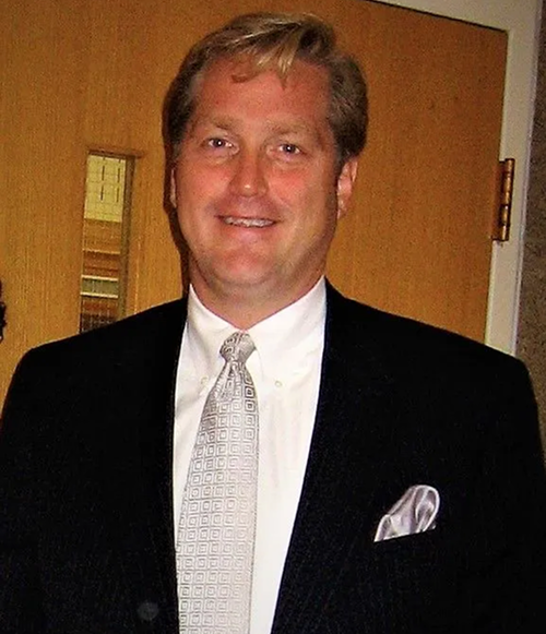 Man in a black suit and white tie smiling, with a pocket square, standing in front of a wooden door.