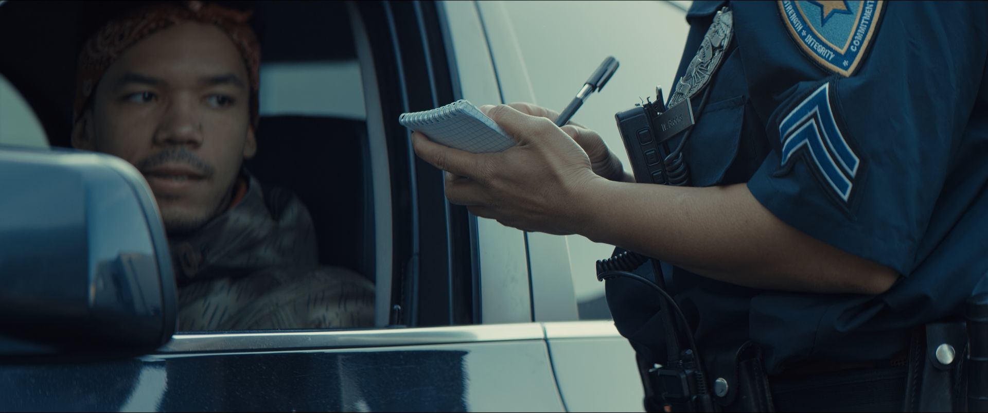 A police officer in uniform stands by the window of a silver sedan, handing a document to the driver on a suburban road.