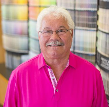 A man wearing glasses and a pink shirt is standing in front of a wall of paint samples.