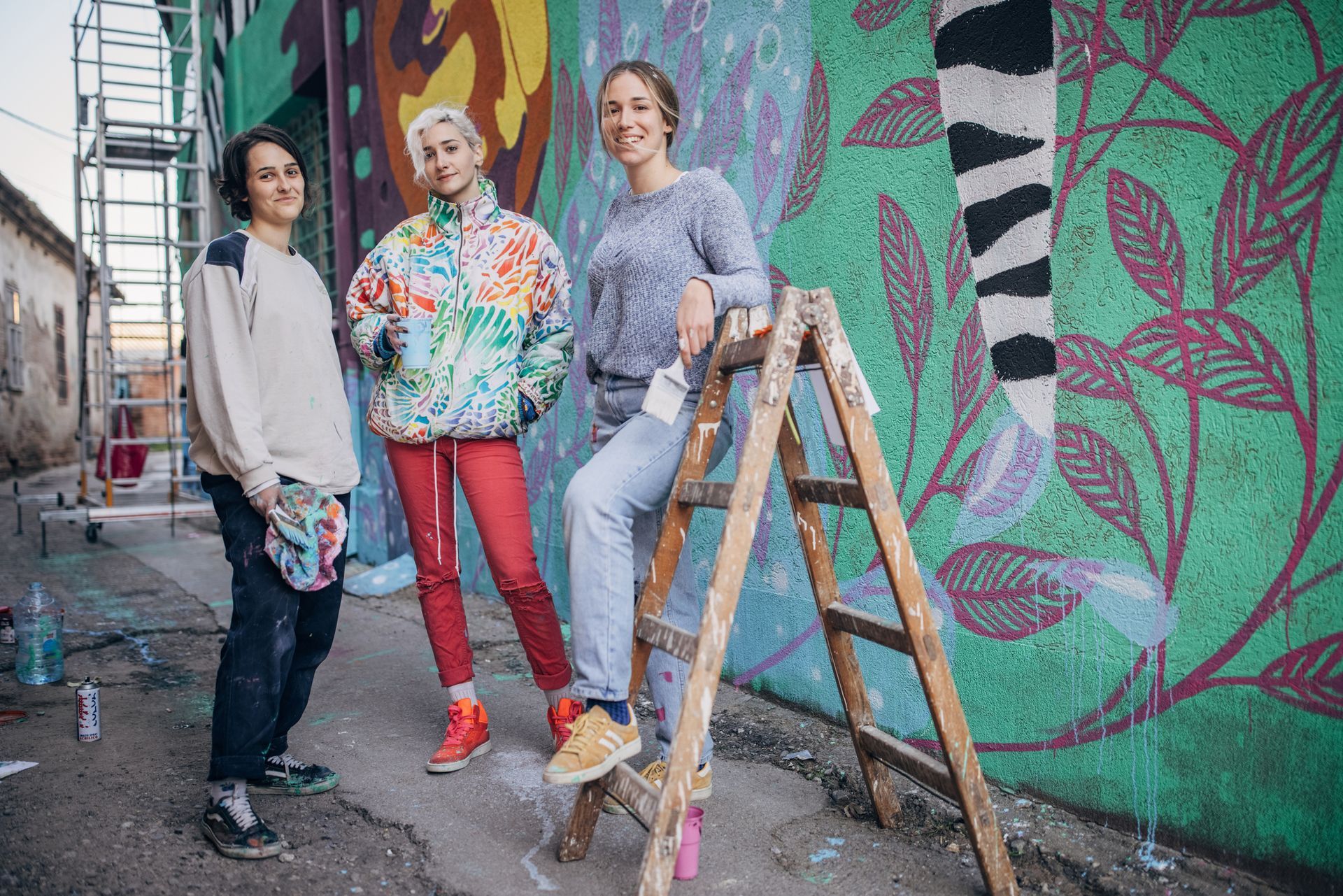 Three women are standing next to each other on a ladder in front of a mural.