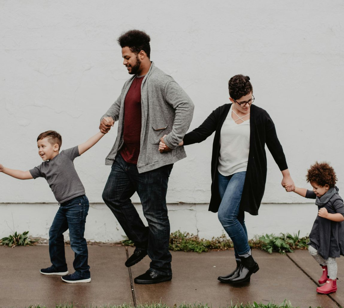 Family holding hands, walking playfully along a sidewalk in front of a white wall.
