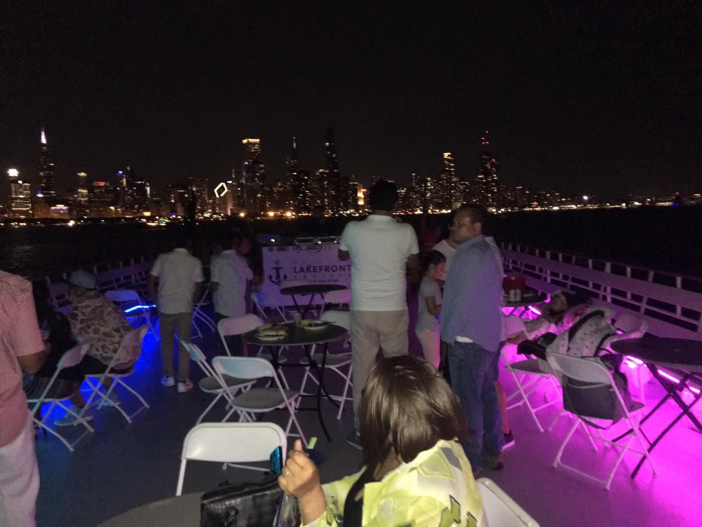 People on a boat deck at night, with purple lights and a city skyline in the background.