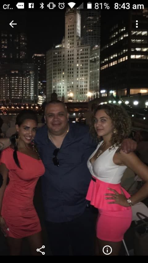 Three people posing at night by a river, with city skyscrapers and lights glowing in the background.