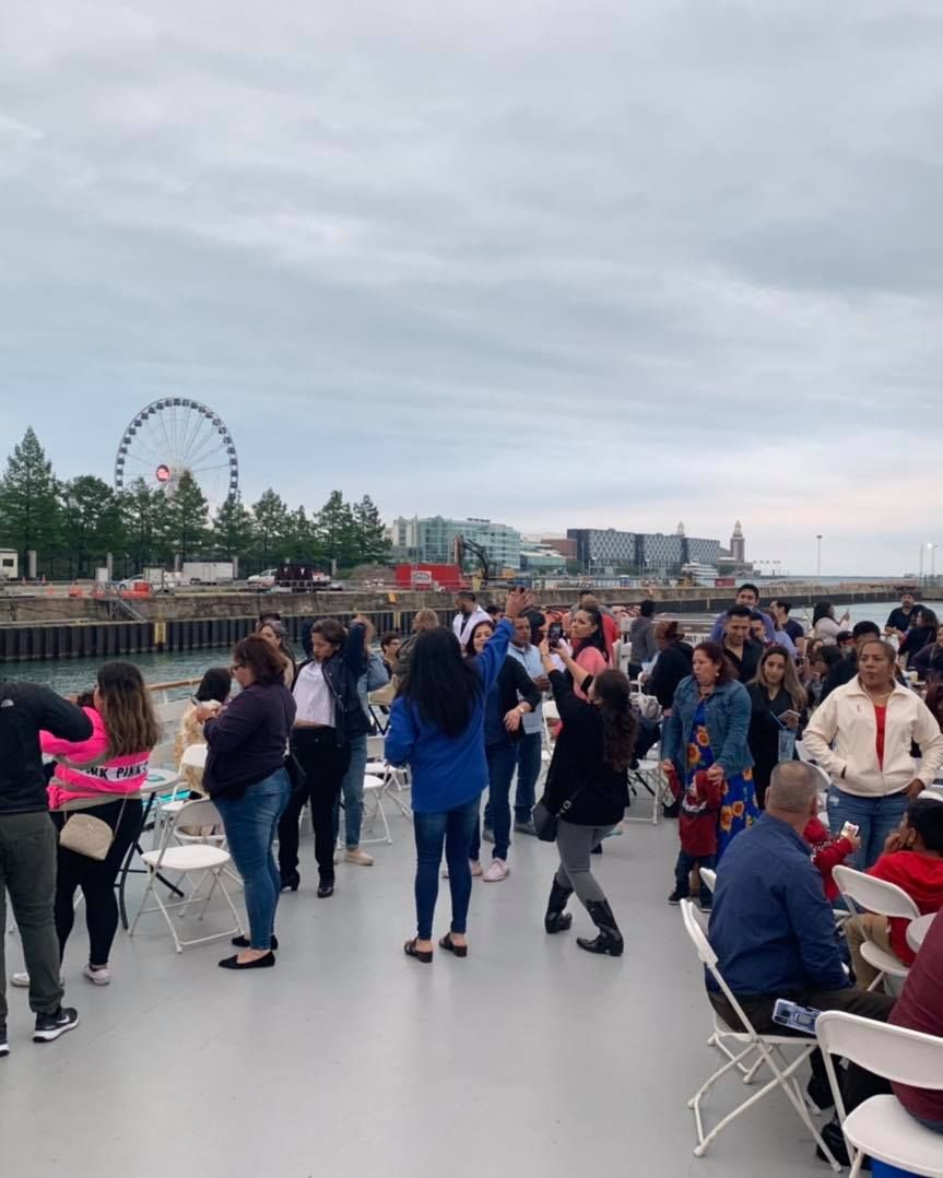 Crowded waterfront boardwalk with people gathering near tables, a Ferris wheel in the distance under cloudy sky