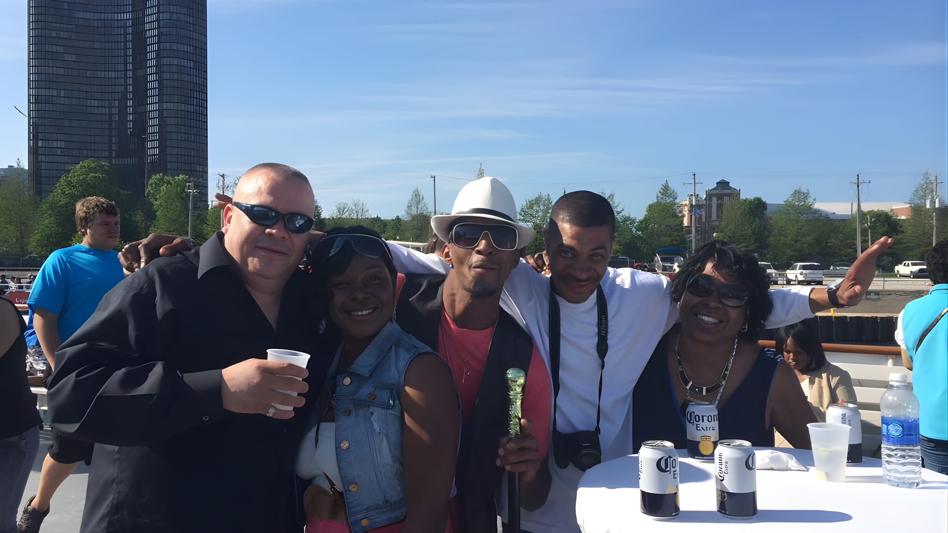 Group of people smiling outdoors at a sunny event, standing by a white table with drinks and a city backdrop