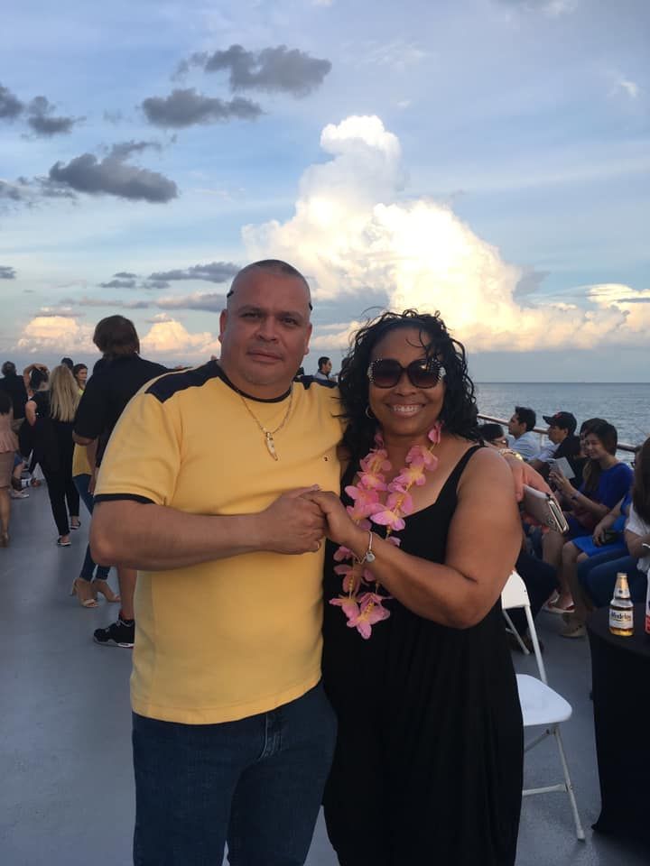 Two people posing together on a waterfront deck at sunset, with a cloudy sky and seated crowd behind them