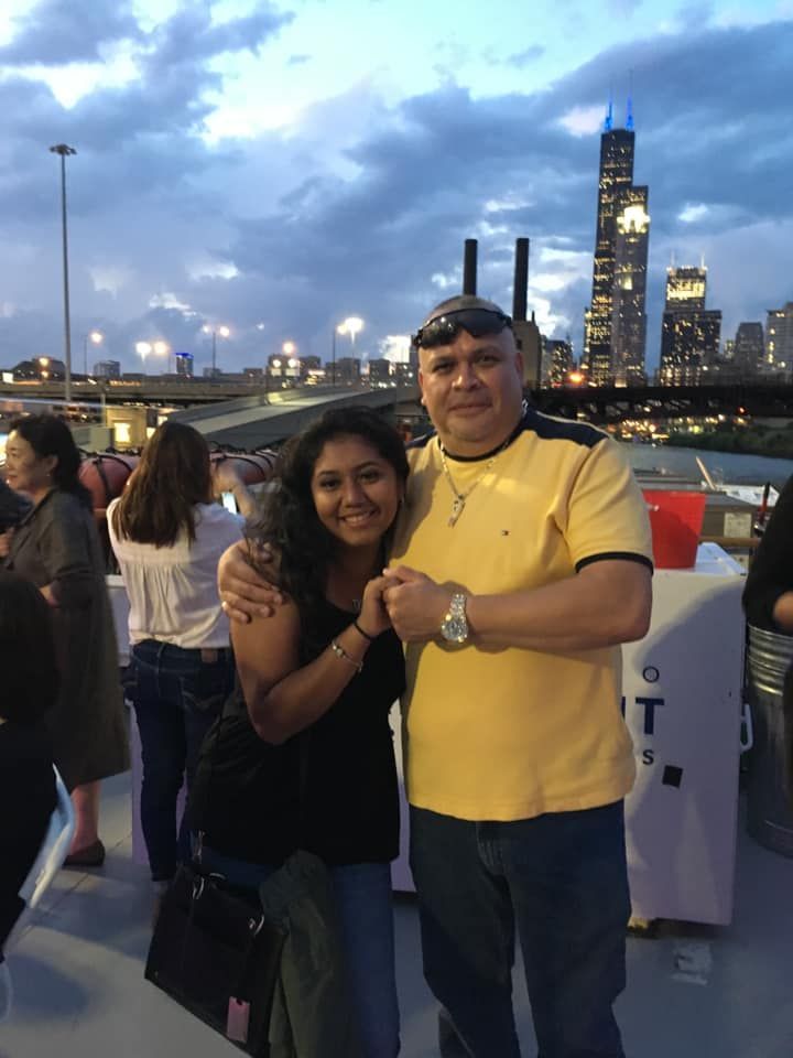 Two people posing on a riverside walkway at dusk with a city skyline glowing in the background.