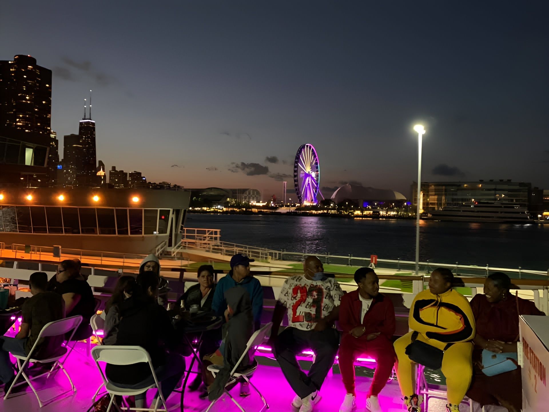 People seated on a ferry deck at dusk, with city lights and a lit Ferris wheel across the water.