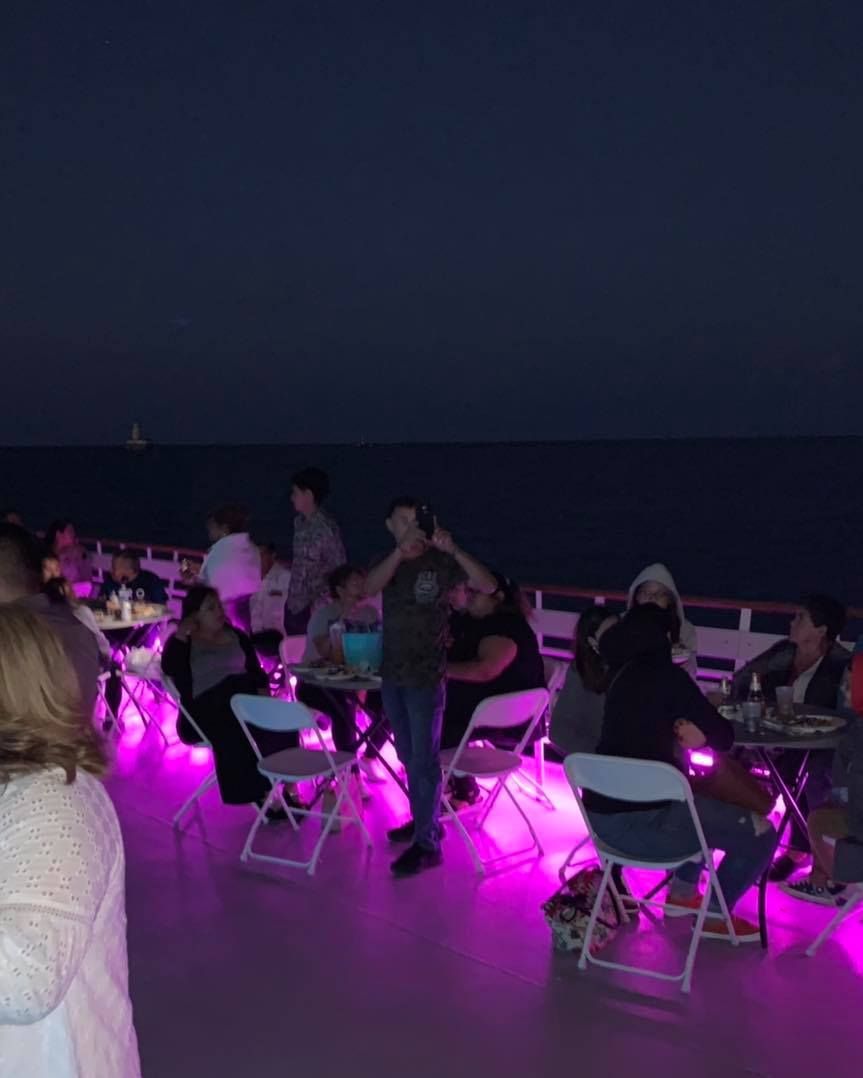 People seated on a dimly lit outdoor deck with pink lights, facing the dark ocean at night.